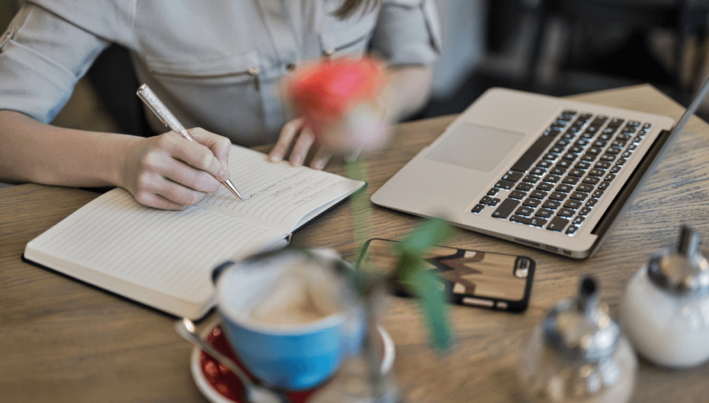 How to Promote Self-Published Books. Person writing in book with laptop. (Flyintobooks.com)