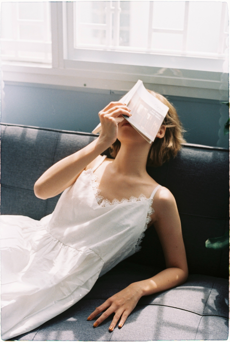 Beautiful girl wearing a white dress, covering her face with a book and laying down on a sofa. Book blog list and book blogger list (Flyintobooks.com)
