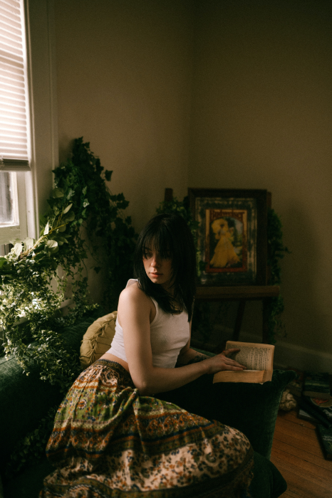 Beautiful girl with dark hair wearing a white singlet and dark red long skirt reading an old book in a dark room with sunlight coming through the window.