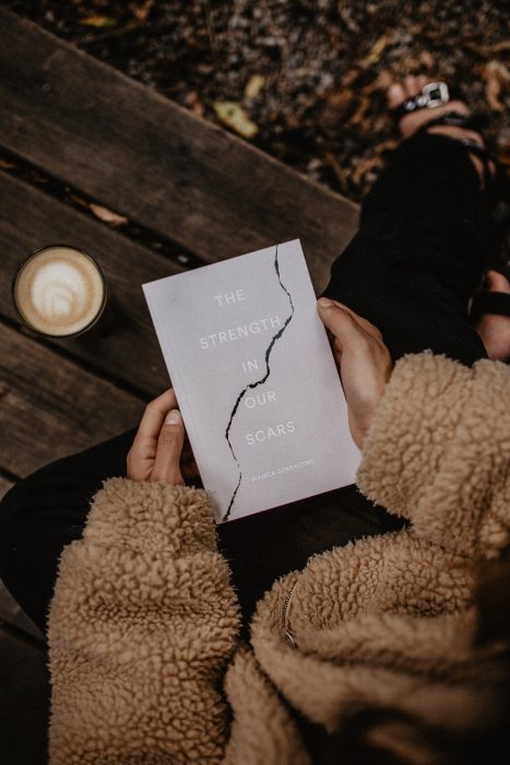 Girl wearing a thick coat holding a book called "the strength in our scars". A latte in a glass in next to her. Book blog list and book blogger list (Flyintobooks.com)