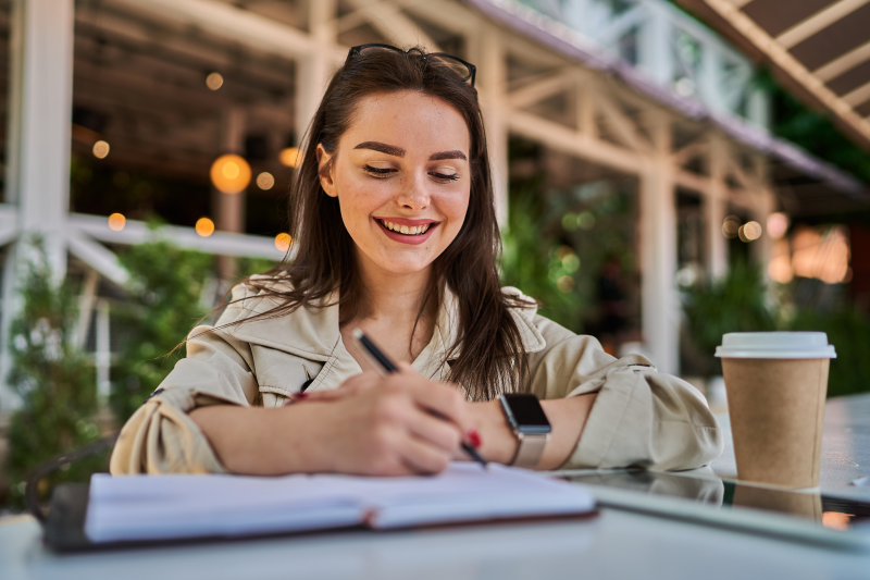 Woman smiling and writing in a notebook with a coffee.  (flyintobooks.com)
